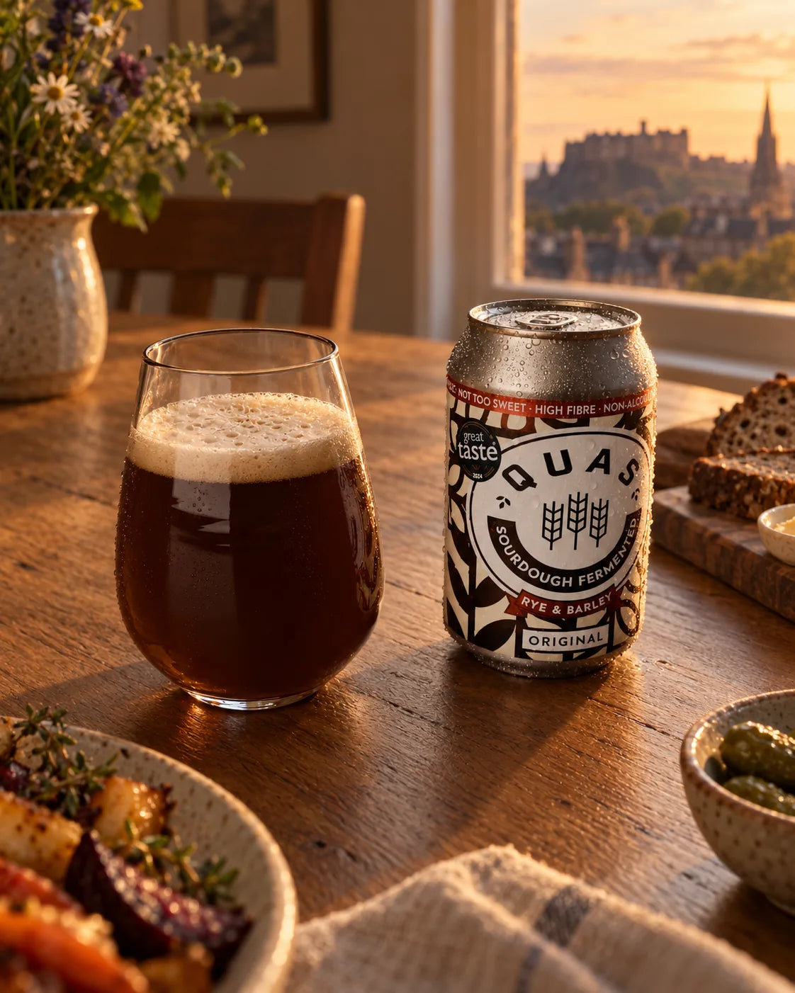 A can of Quas Original beside a tulip glass of cloudy, dark Quas on a dinner table with rye bread and roast vegetables, and an Edinburgh skyline through the window.
