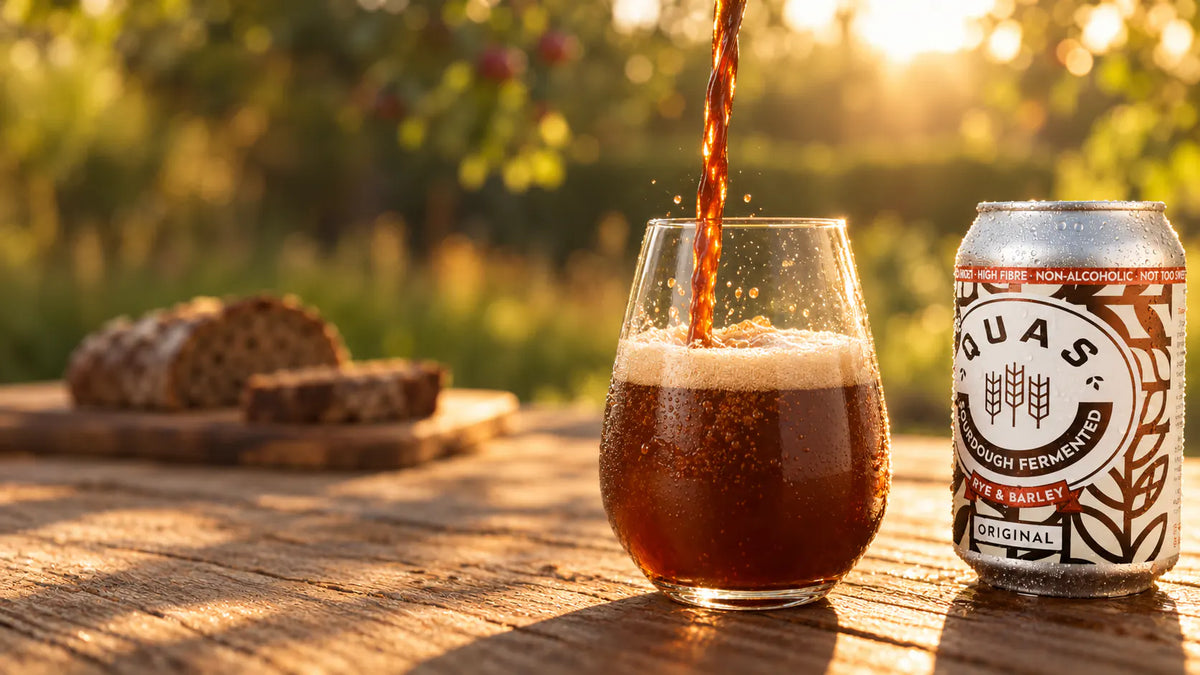 Quas being poured into a stemless glass on a garden table in low evening sun, beside a cold can of Original Quas and a loaf of rye bread