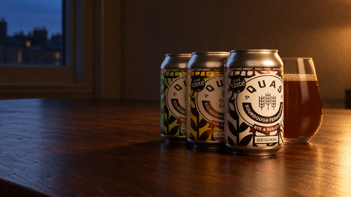 Quas Mint & Lime, Ginger and Original cans beside a poured glass of Quas on a walnut table in lamplight at dusk