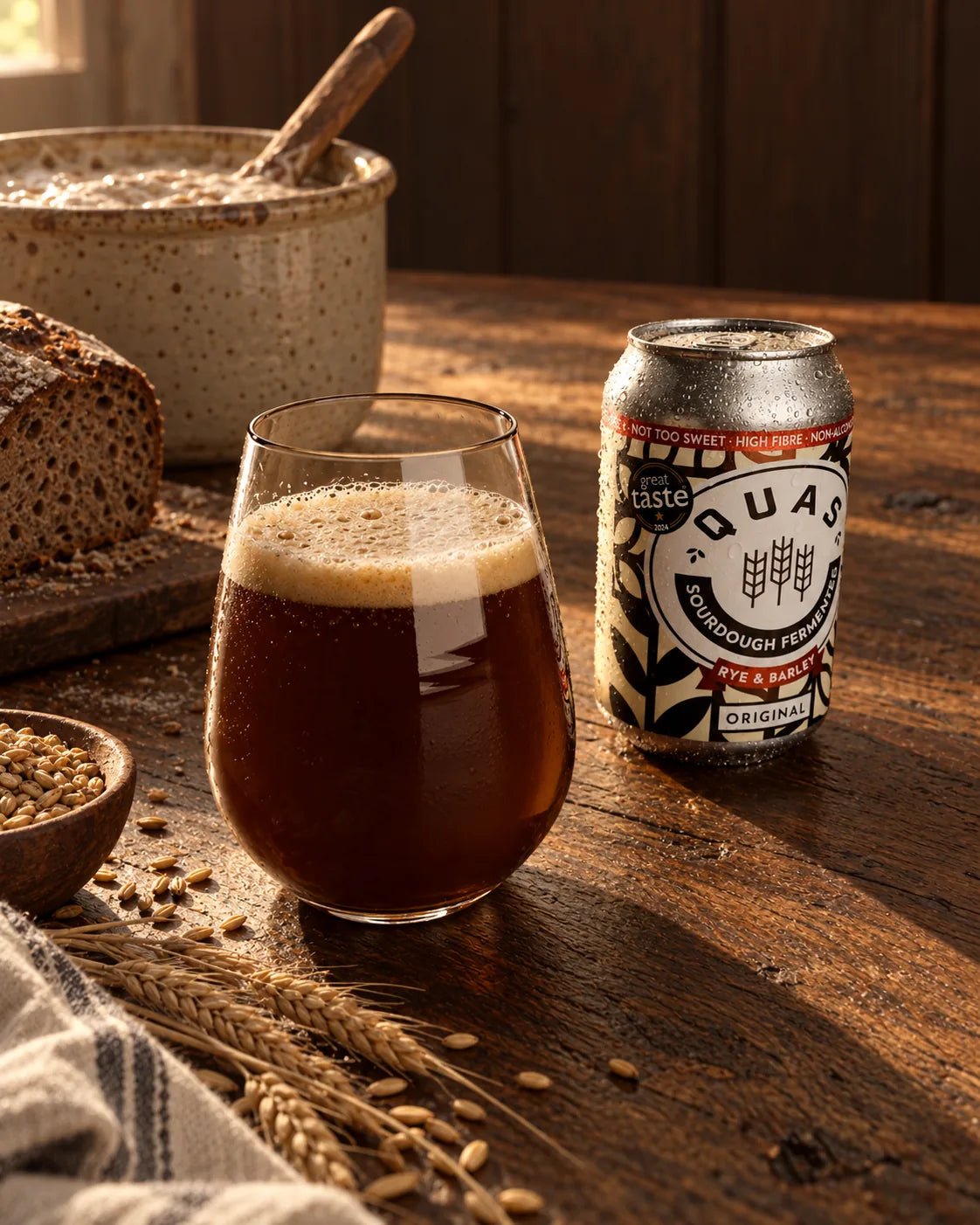 A can of Original Live Quas beside a glass of cloudy, dark kvass, with rye, barley and a sourdough crock on an oak table