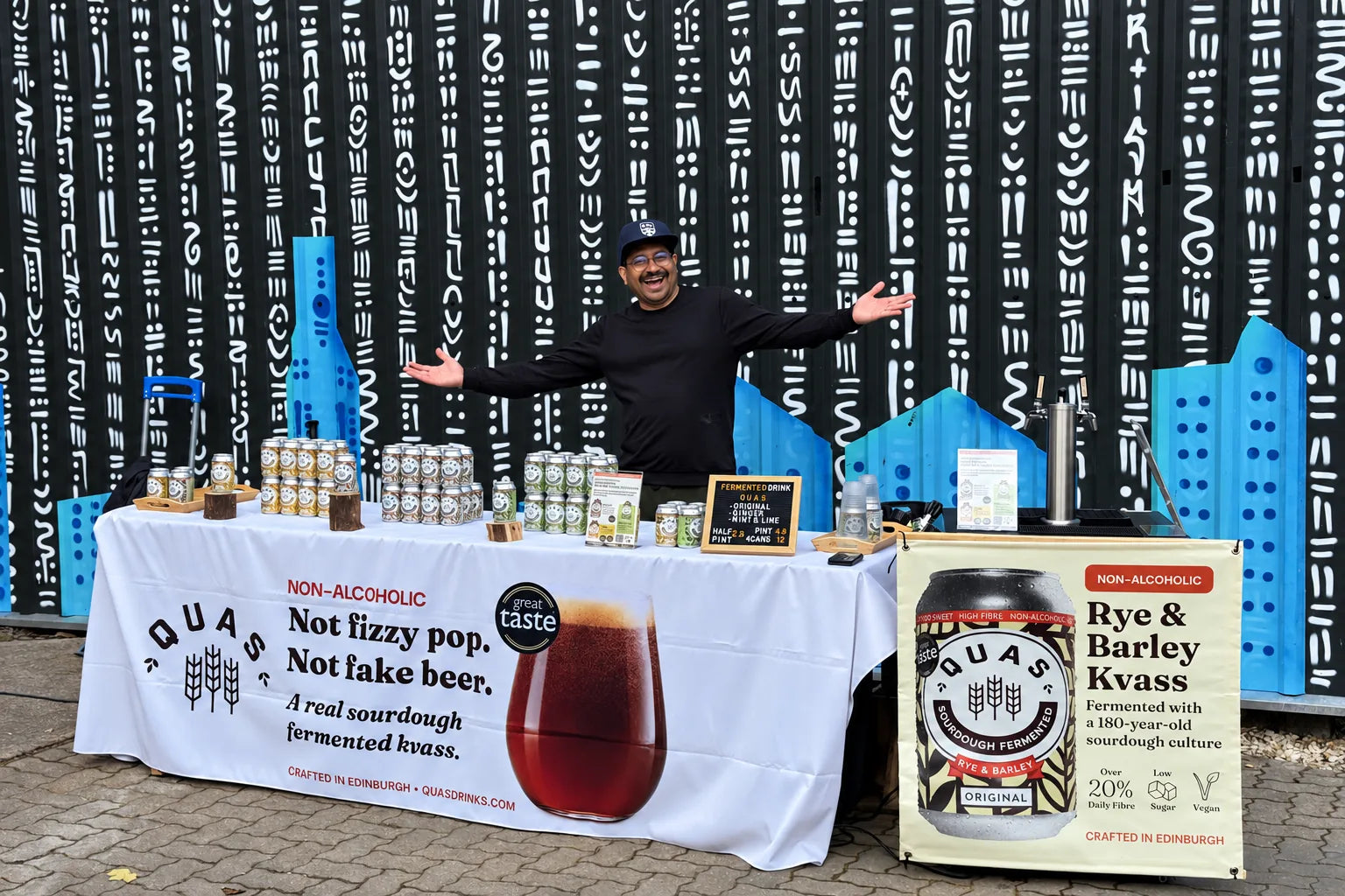 The Quas stall at Soul Gathering: a team member welcomes visitors behind a table of Quas cans, a tap and Rye & Barley Kvass banners, in front of a painted wall.