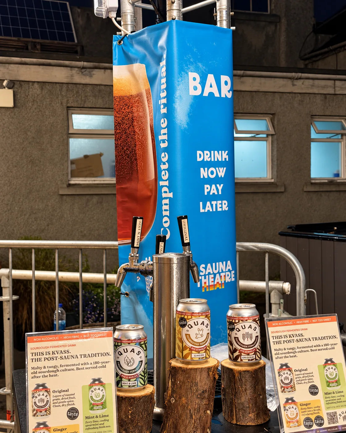 Quas taps and cans of Original, Ginger and Mint & Lime on log stands at the Sauna Theatre bar, under its Complete the ritual banner.
